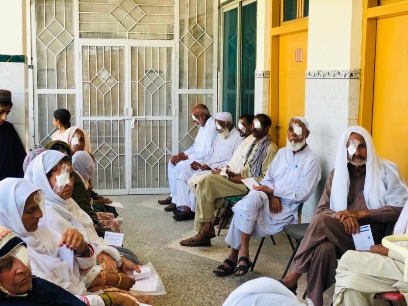 Elderly men and women seated in a corridor with dressings over one eye after cataract surgery.
