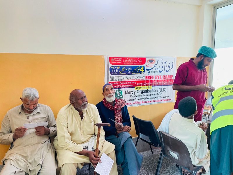 Elderly men seated on chairs waiting beside a Free Eye Camp banner.