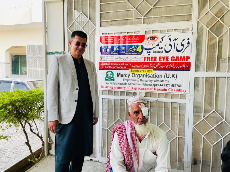 An elderly bearded man in a red and white scarf with an eye dressing, sitting outside the clinic.