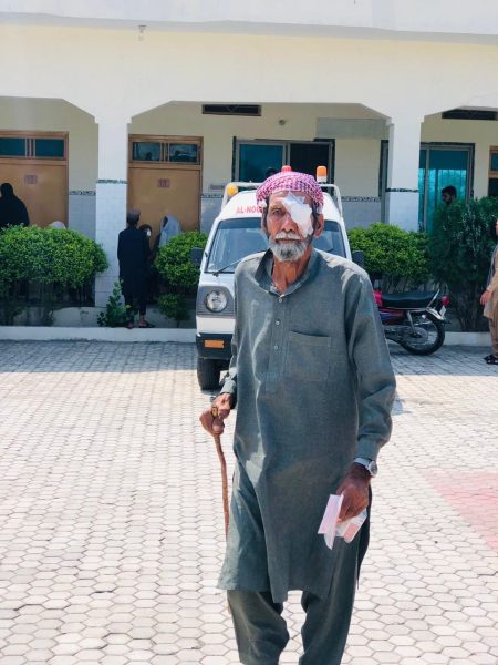 An elderly man with an eye dressing and a walking stick standing in a hospital courtyard.