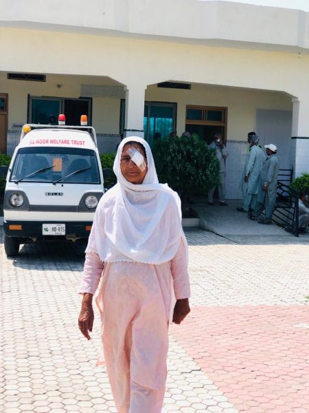 A woman in a white headscarf with an eye dressing walking across a hospital courtyard.
