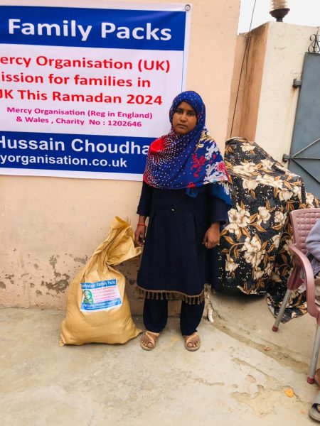 A woman standing beside a food sack under a Mercy Organisation banner.