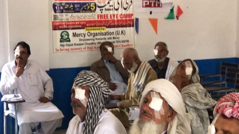 Elderly patients with eye dressings seated beneath a Free Eye Camp banner.