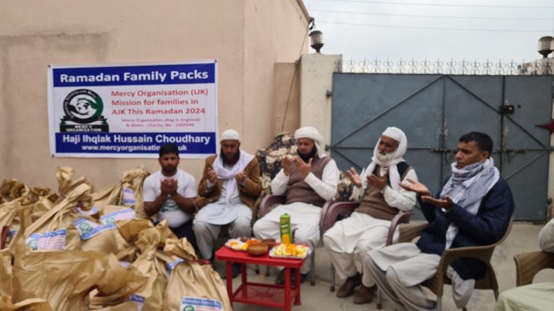 Men seated in a courtyard beside piles of food packs and a Ramadan Family Packs banner.
