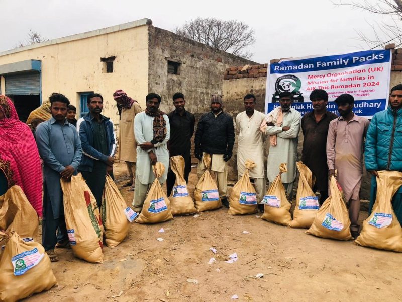 Men standing behind a long row of filled food sacks beside a Mercy Organisation Ramadan Family Packs banner.