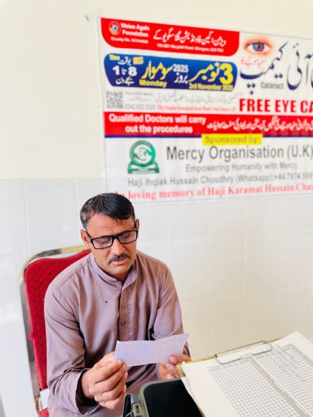 A man filling in a registration form at a desk beside a Free Eye Camp poster.