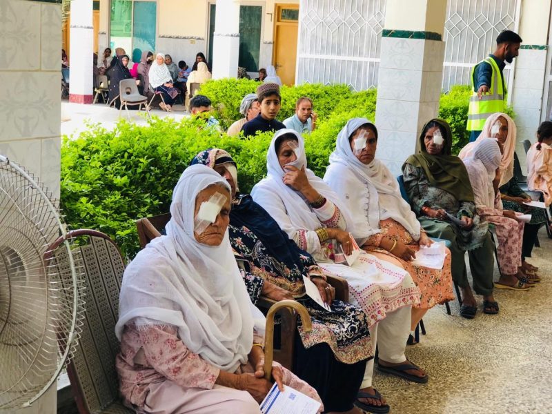 A row of women with eye dressings sitting outside a hospital ward after cataract surgery.